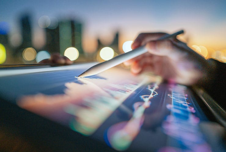 Close-up of a hand holding a stylus, interacting with a tablet displaying colorful graphs. The background features blurry city lights at dusk.