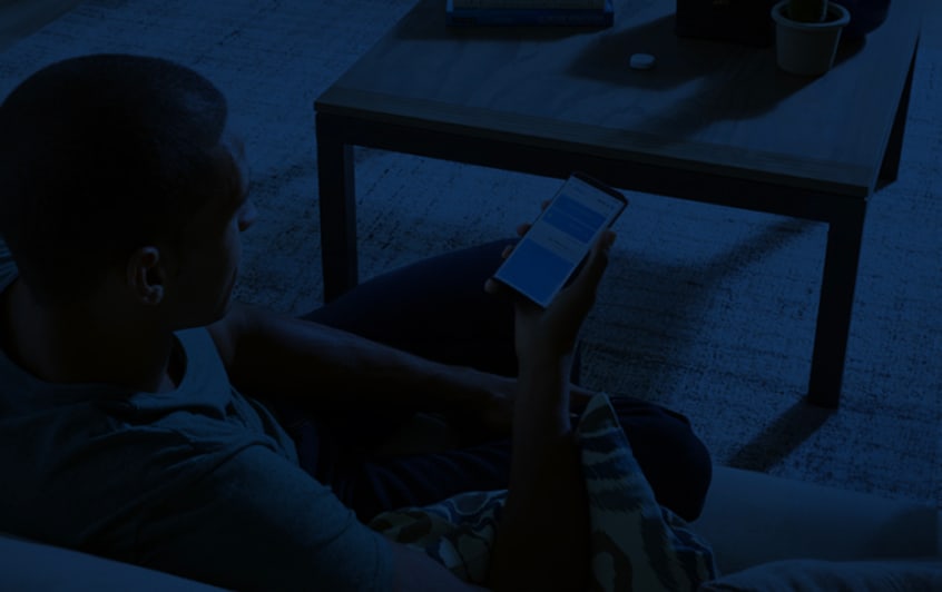 Una persona sentada en un sofá, mirando la pantalla de un smartphone en una habitación tenuemente iluminada, con una mesa baja de madera cerca.