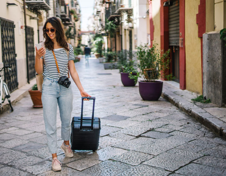 woman walking and pulling a rolling suitcase while looking at their phone