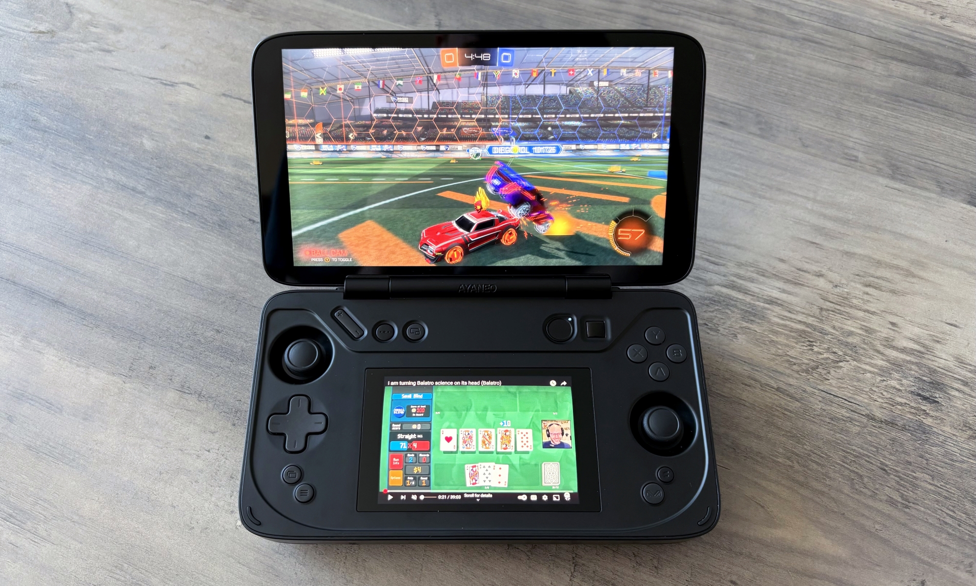 The Ayaneo Flip DS gaming handheld rests on a light brown wooden table, with its top screen showcasing the game Rocket League and its bottom screen playing a YouTube video.