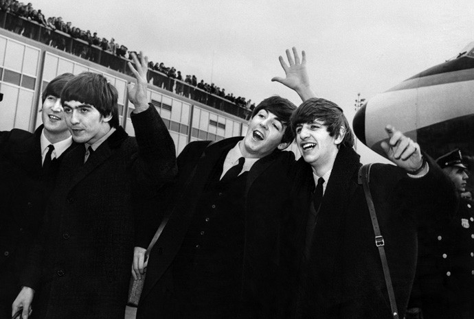 John Lennon, Ringo Starr, Paul McCartney e George Harrison desembarcam no aeroporto John F. Kennedy Airport, em Nova York, EUA, onde são recebidos por multidão de fãs em fevereiro de 1964