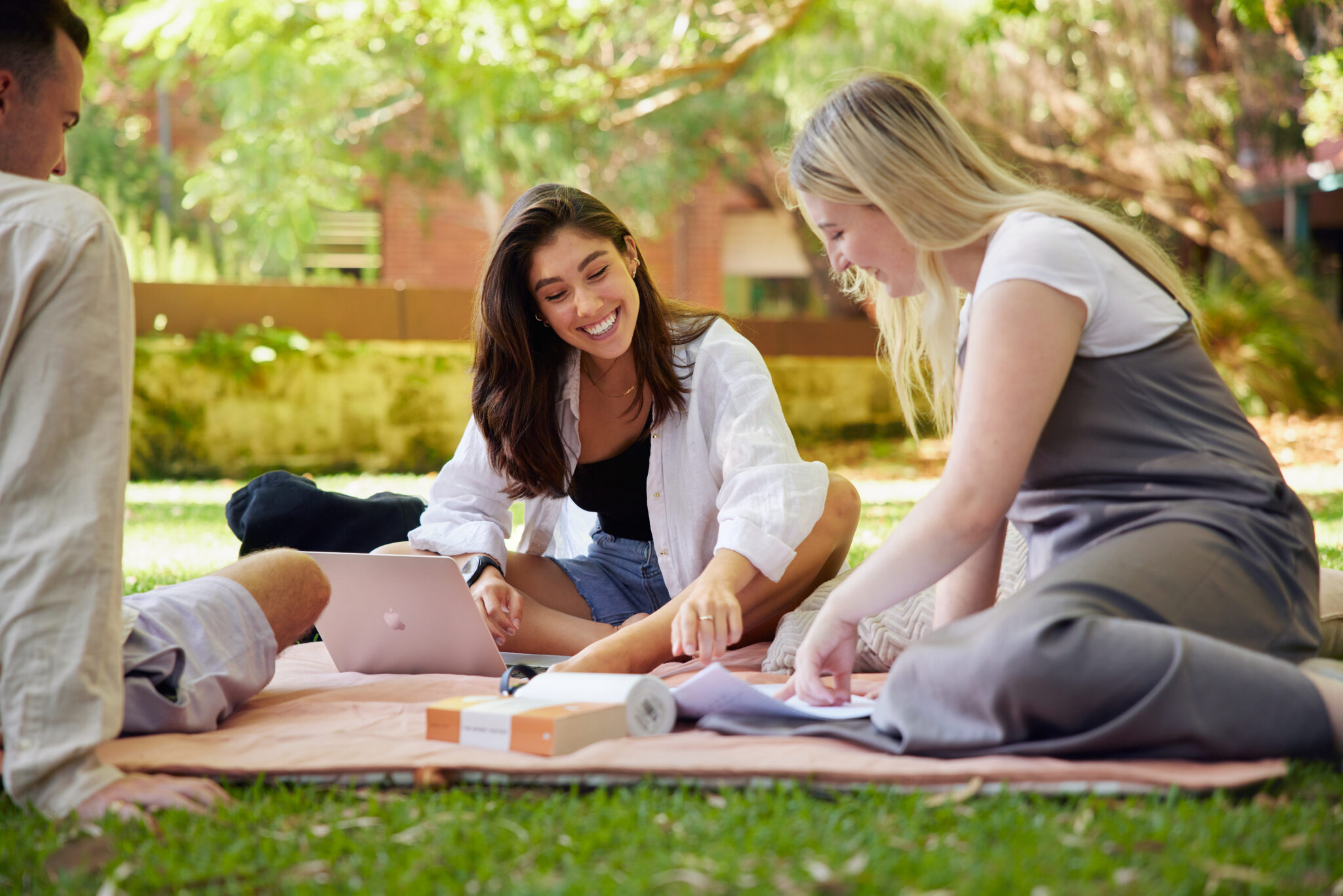 Two young women sit on a picnic blanket smiling and looking at a page of notes with an open laptop in front of them; a young male is also sits on the blanket on the edge of the image.