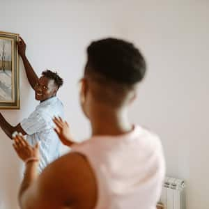 Young Black man hangs a picture on a wall while his partner tells him if it’s straight 