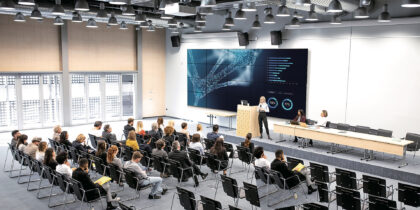 An auditorium with about two dozen seated trainees, listening to a speaker on stage in front of a large TV monitor.