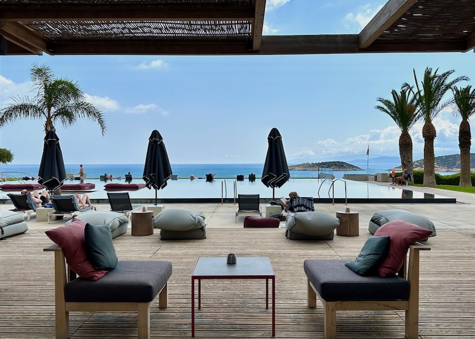 Infinity pool on the sea surrounded by a wooden sun deck with jewel-toned furniture and palm trees at Minos Palace resort in Crete