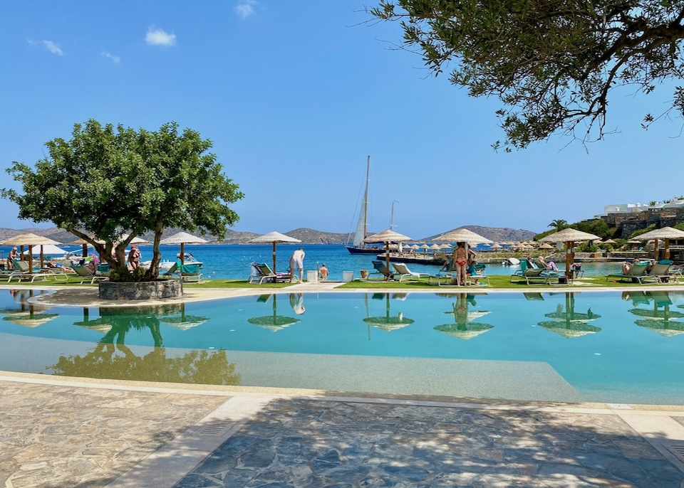 Freeform pool set behind a beach with sunbeds and umbrellas on the side and sailboats in the distance at Porto Elounda hotel in Crete