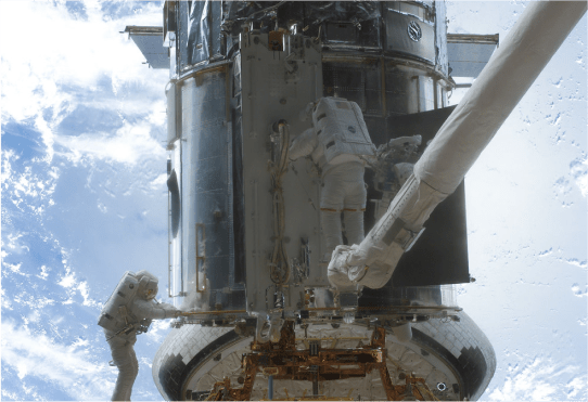 Two astronauts working on Hubble while in space. One astronaut is on the left, holding on to the lower handrail of Hubble. The astronaut on the right is standing on top of the CANADARM, a long robotic arm, he works on the center bottom of Hubble. In the distance behind Hubble, the blue Earth is visible with wisps of white clouds.