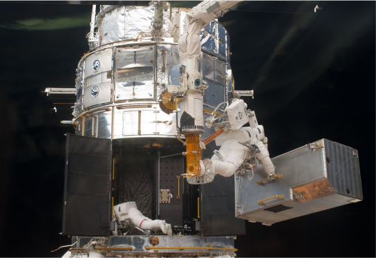 Two Hubble astronauts working during Servicing Mission 4. One of the astronauts is carrying a large silver box, about the size of a telephone booth. He is standing on the long robotic CANADARM, to his left there is another astronaut, sideways and working inside Hubble, which has two giant bay doors open at the bottom.