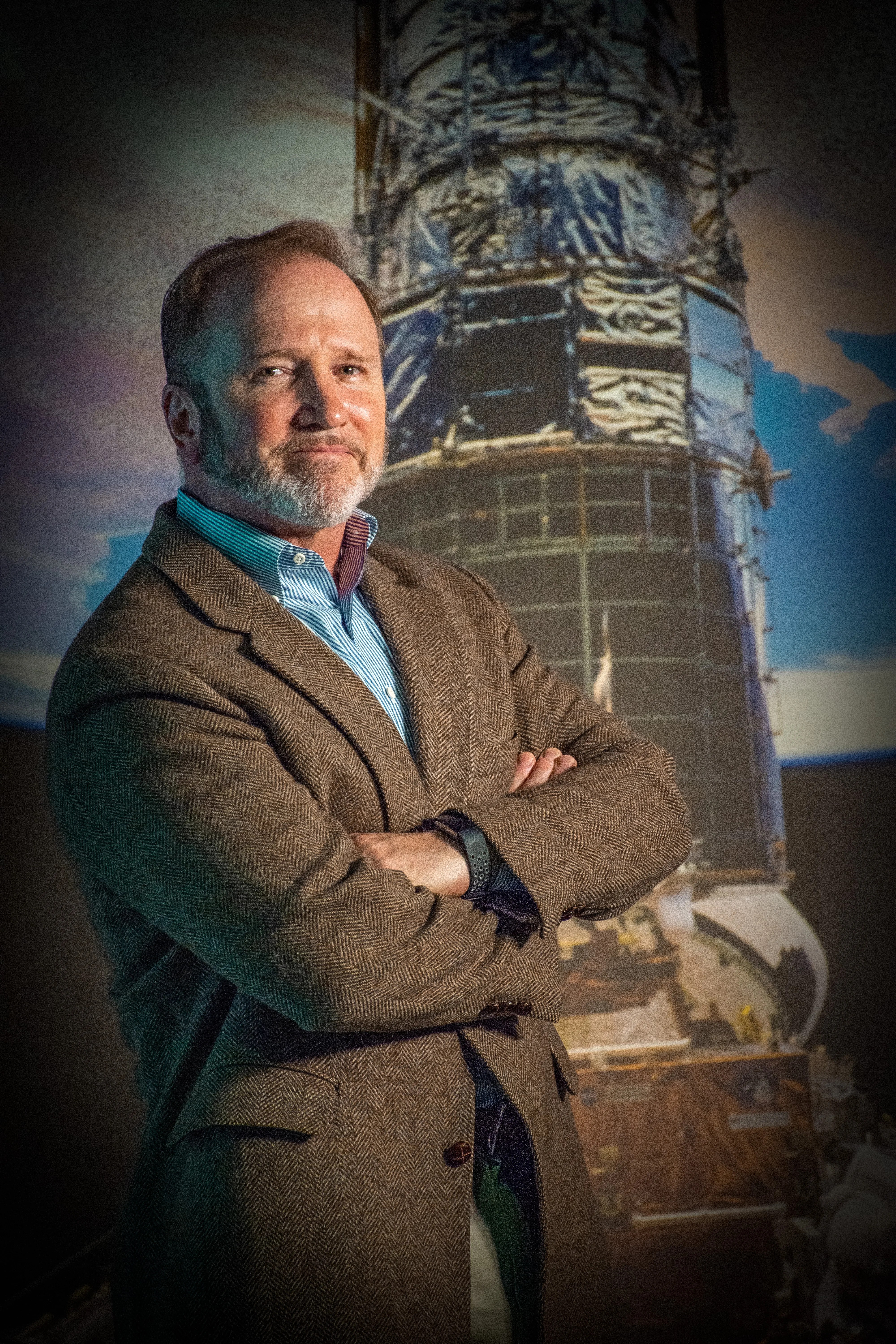 Dr. Richard Barry wearing a blue shirt and brown suit stands with arms crossed in front of a background photo of Hubble.