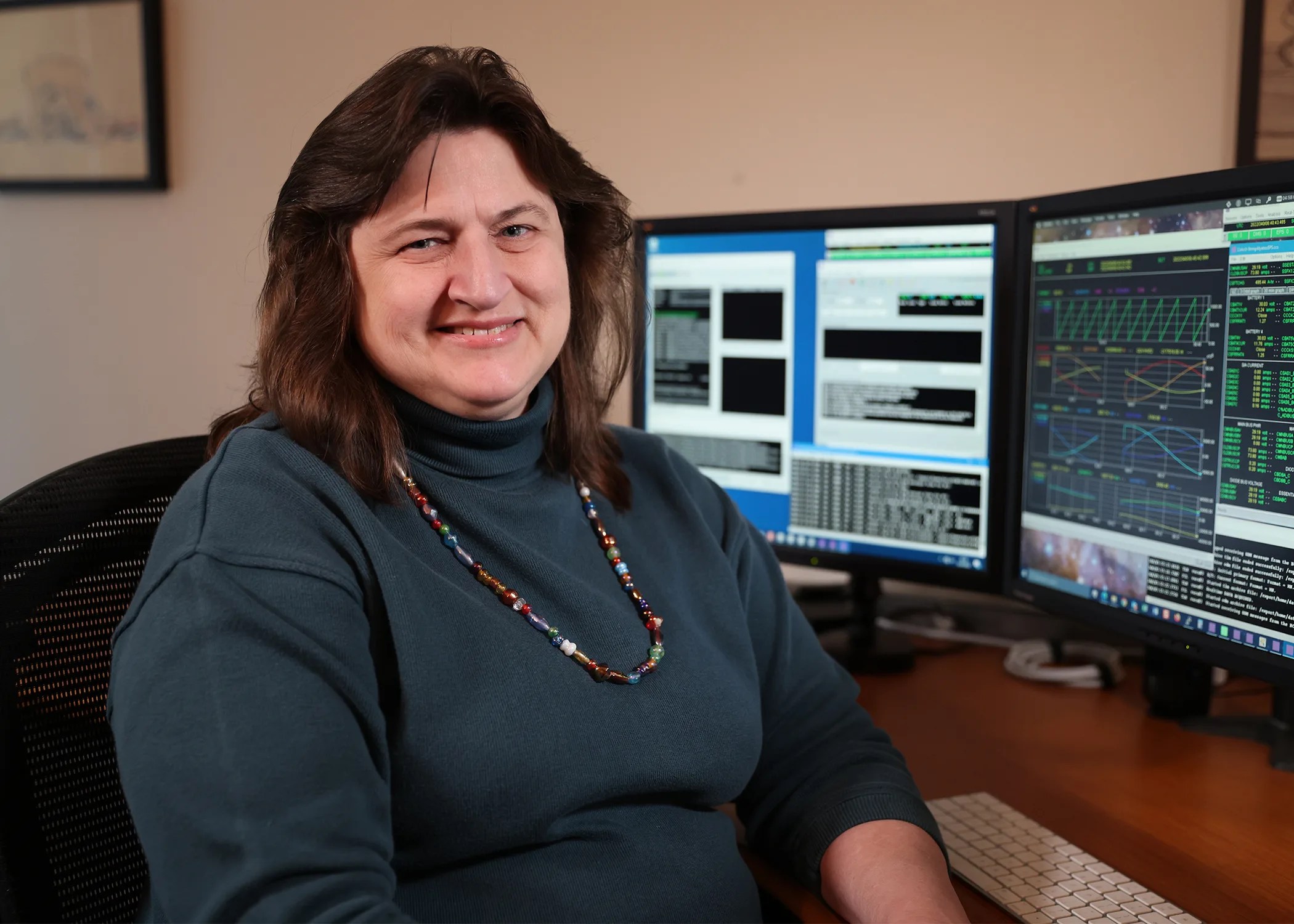 headshot of woman with brown hair wearing a necklace and dark green shirt sitting at a desk in front of two computer monitors