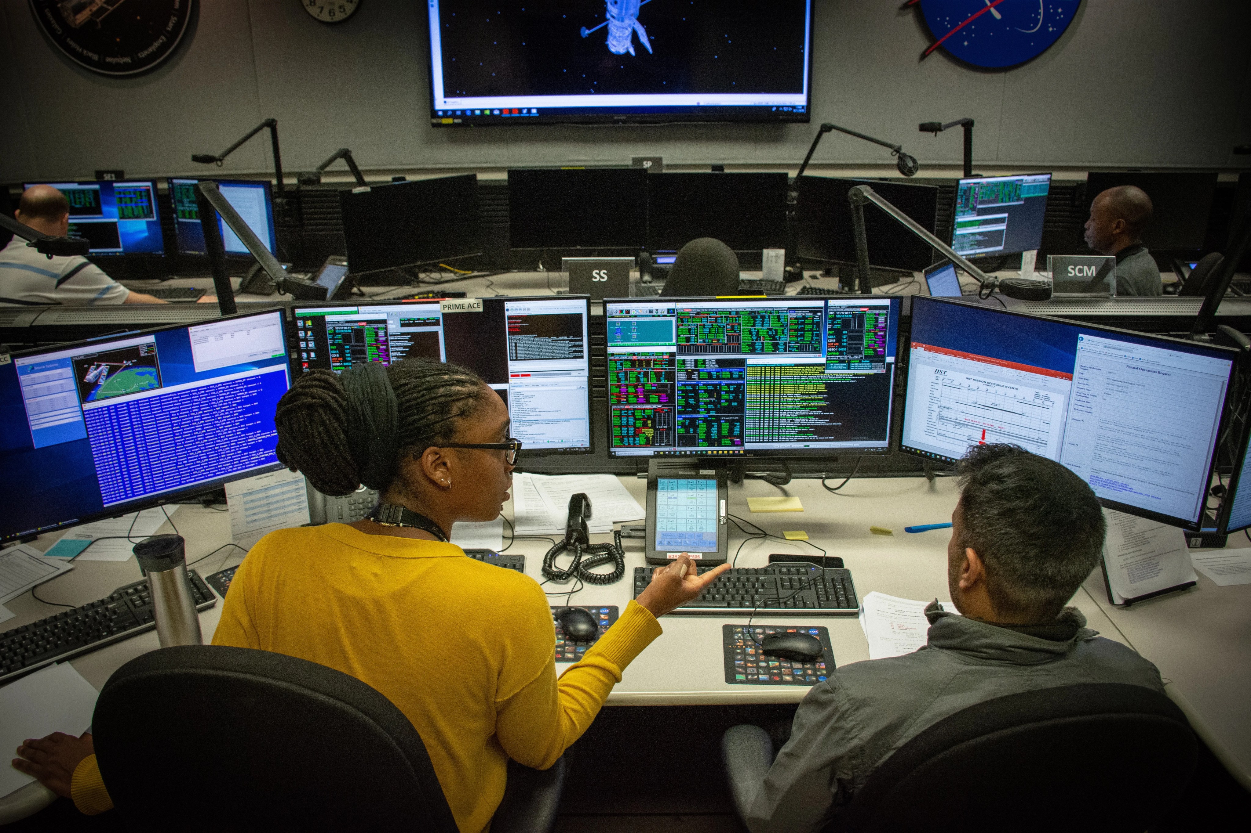 woman and a man sitting at a desk with multiple computer monitors, the woman is pointing at a monitor