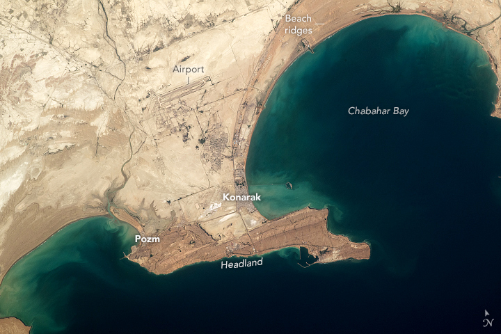 A coastline from upper right to lower left with two large bay erosion regions interrupted by a protruding line of land. The land is entirely beige and sandy, and the water is a light blue-green shade along the coast, especially within the bays, but very dark blue otherwise.