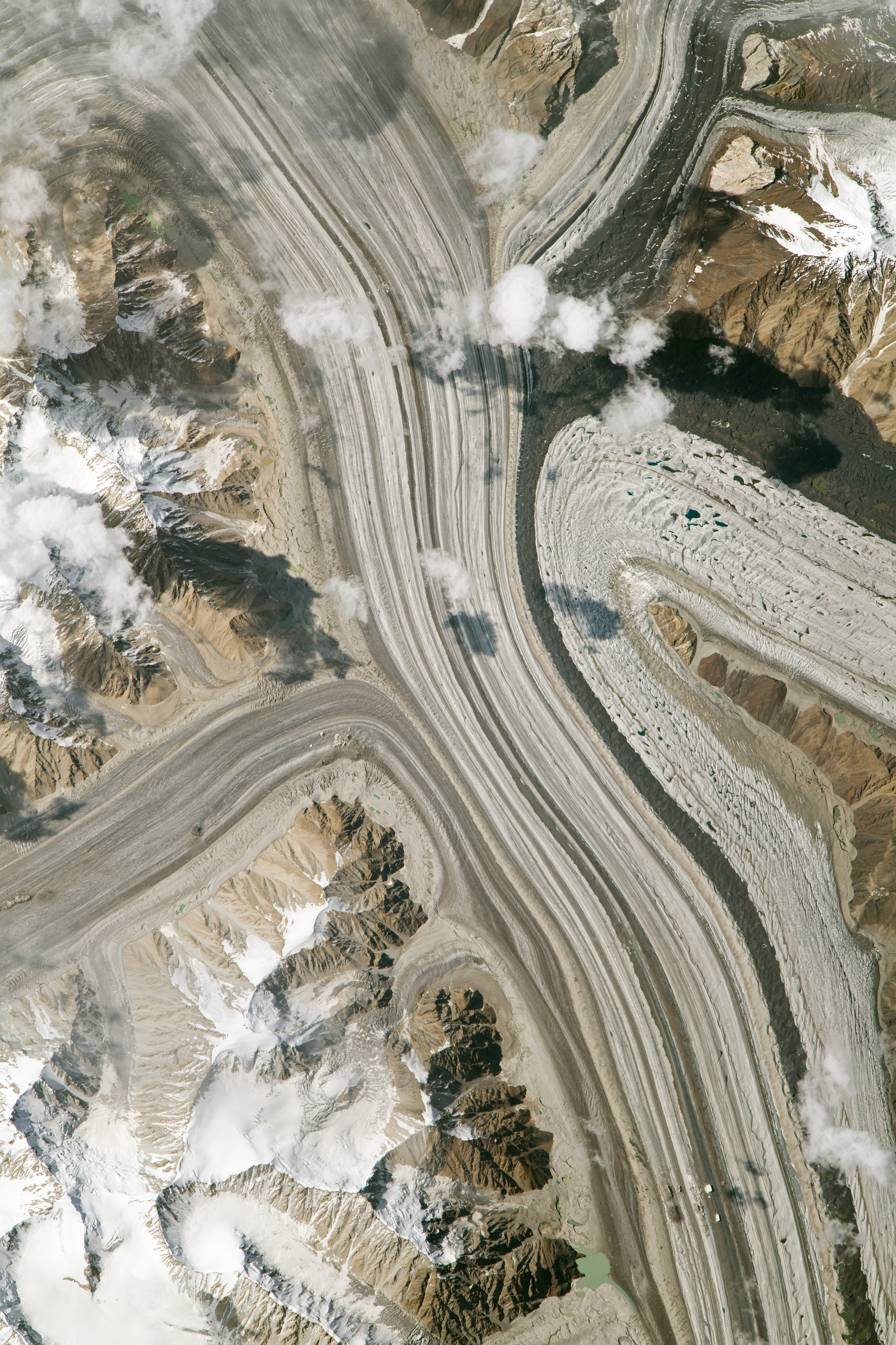 The image shows four smaller tributary glaciers flowing into the main Siachen Glacier. The many dark, parallel lines in the glacier and its tributaries are moraines, which give a sense of the southward flow of the glacier. The darkest and most prominent moraine results from the merging of moraines from the Teram Shehr Glacier and its neighbor immediately to the north. Early afternoon sunlight casts shadows from small cumulus clouds onto the glaciers’ surfaces.