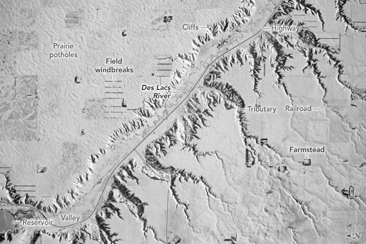 A completely snow covered landscape shows signs of the landscape below. A river valley traverses upper right to the bottom left corner of the image. To the right of the river valley there are numerous gullies with tributaries that feed into the river. To the left is a more flat open field.