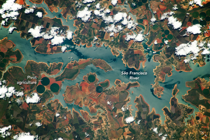 The light blue green river has dozens of peninsulas with orange borders between the land and water. The land has circular crop fields and a lot of rust red/orange ground as well as dark green vegetation.