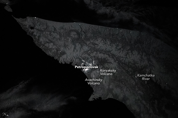 In this grayscale image the valley broadens from a narrow point in the top left to a cone that spans the center of the right edge to the lower bottom right. The lighting of a town can be vividly seen as white against the backdrop of craggy landscape filled with valleys leading to the low center where the Kamchatka river appears completely dark. The land surrounding this valley seems to be completely blacked out.