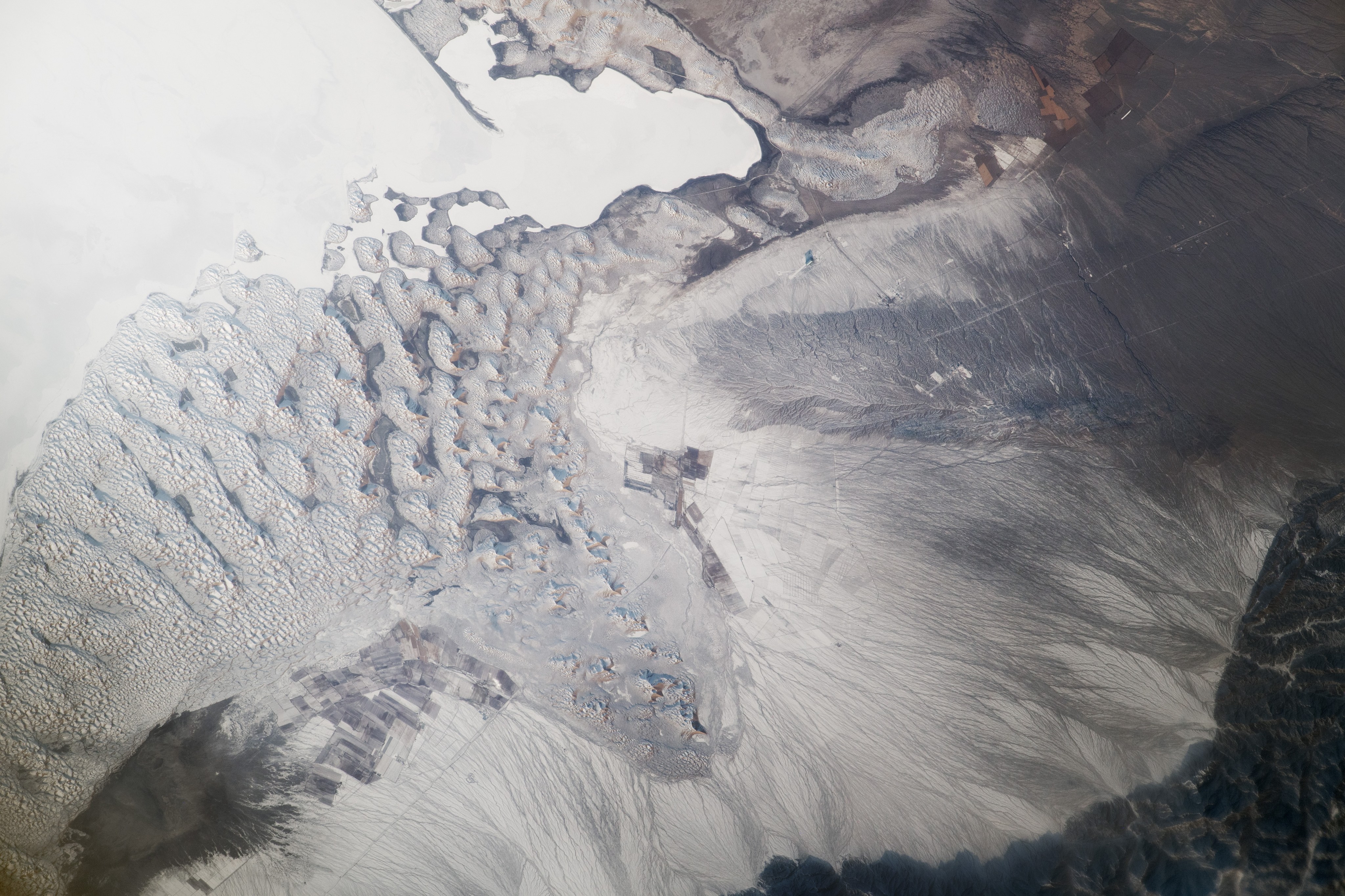 The featureless area at the top left is the smooth water surface of Bositeng Lake, a large lake in the far northwest of China. The slightly curved sand spit offers one indication that the monotonous area is a lake. Spits such as these are common features near lake shores and are produced by waves and longshore currents. In contrast to the smooth surface of the lake is the intricate pattern of a dune field on its south side. Darker surfaces next to and between some of the dunes are wet.