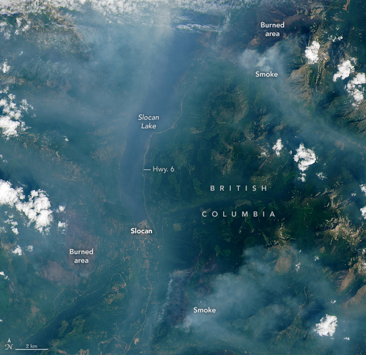 The image shows the valley—bounded by the Valhalla Range to the west and the Slocan Range to the east—filled with smoke. Slocan Lake appears near center, stretching vertically, as a dark poorly defined region through the haze.