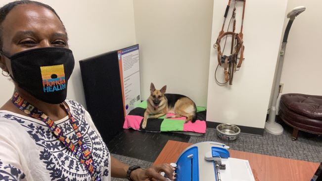 Selfie of woman working at her desk, with dog resting on bed in the background