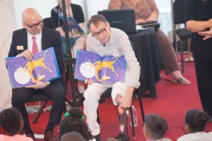 Two men reading from a book titled Giraffes Can’t Dance by Giles Andreae and illustrator Guy Parker-Rees about Gerald the Giraffe who did not dance like the rest of the animals. 