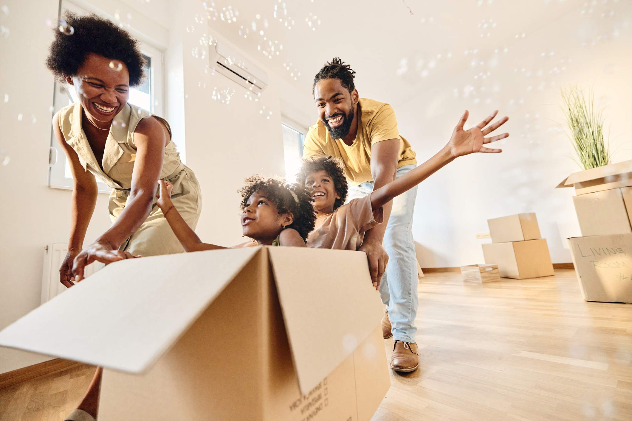 Playful African American parents having fun while pushing their small kids in carboard box at new apartment.