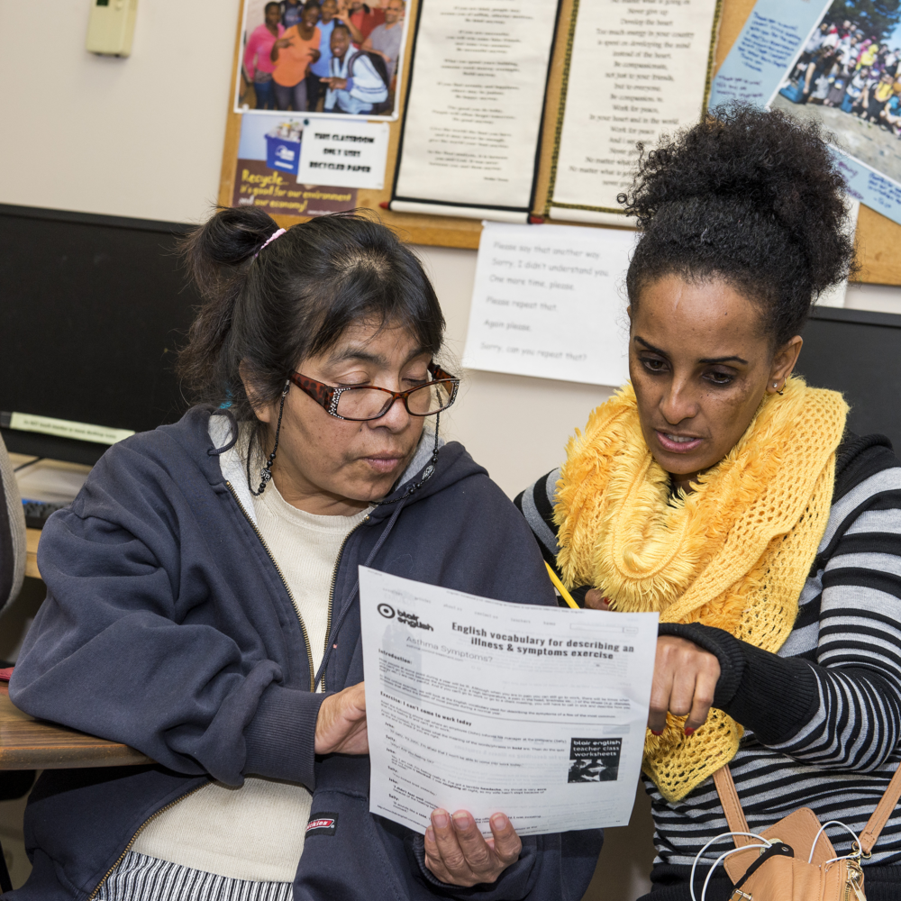 Two women sitting together in a classroom, one pointing to a section on a paper titled "English vocabulary for describing an illness & symptom exercise," as they discuss its contents.