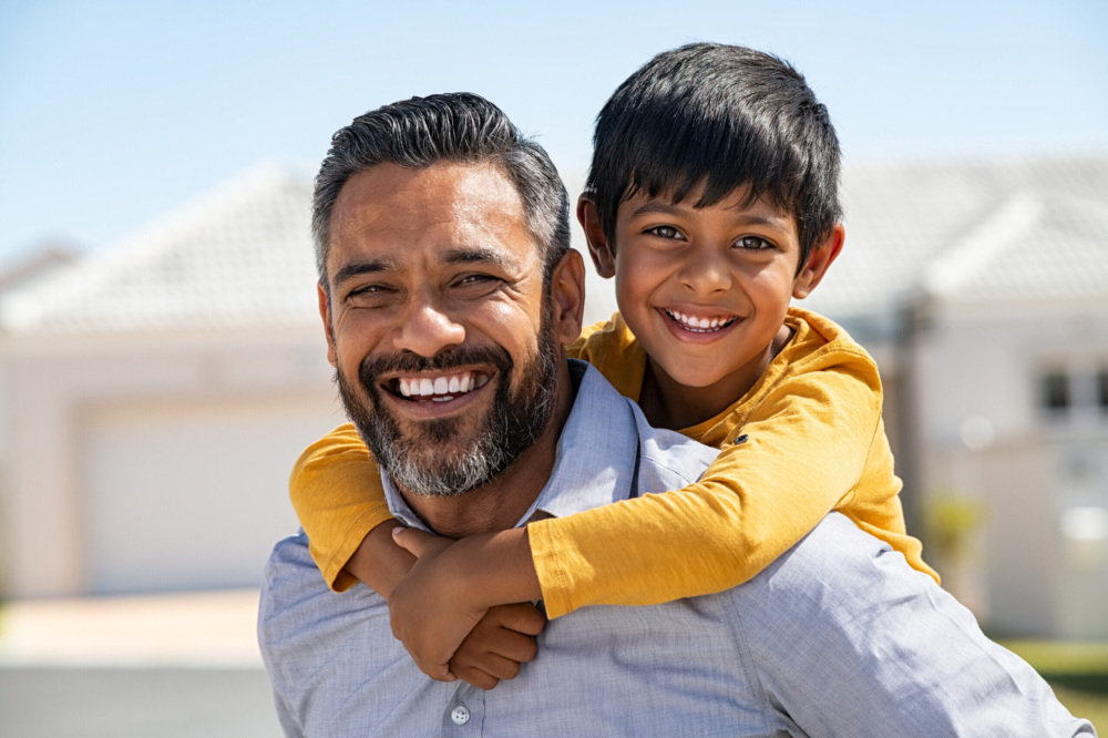 Man with a beard smiling, carrying a child on his back. Both are outdoors with houses in the background. The child is wearing a yellow shirt and also smiling.