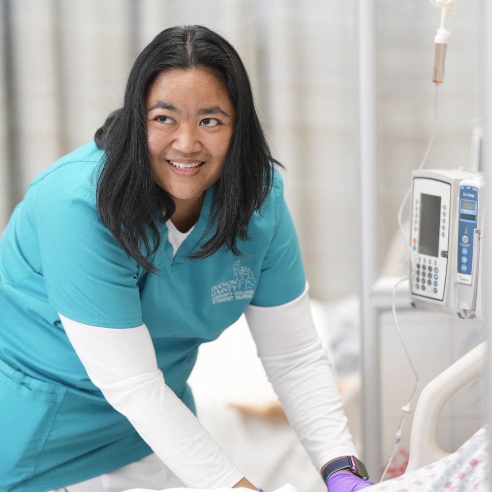 Nurse wearing a teal uniform provides care to a patient, standing next to medical equipment and an IV drip pole.