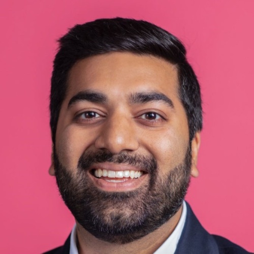 A man with dark hair and a beard is smiling in front of a pink background. He is wearing a black jacket and white shirt.