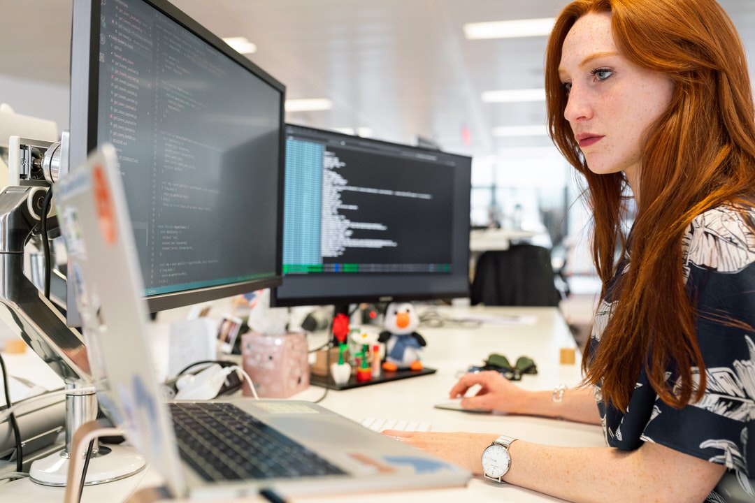 a red head woman sitting at a computer desk with three monitors