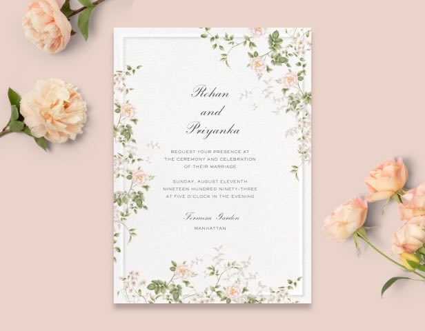 A white invitation with a pink and green floral border on a pink backdrop with pink flowers.