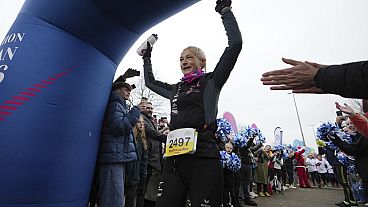 A belga ultrafutó Hilde Dosogne, középen, szurkolnak neki, amikor átlépi a célvonalat a 366. egymást követő maratonján a belgiumi Gentben, 2024. december 31-én, kedden.