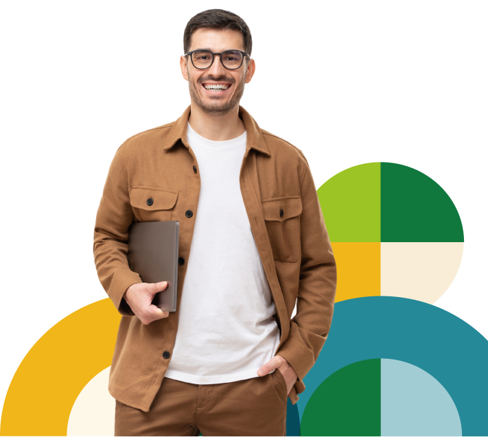 Homem branco de barba usando óculos sorrindo, vestido com casaco e calça, na cor marrom, com camiseta branca. Com uma mão, ele segura o notebook e a outra mão está no bolso da calça.