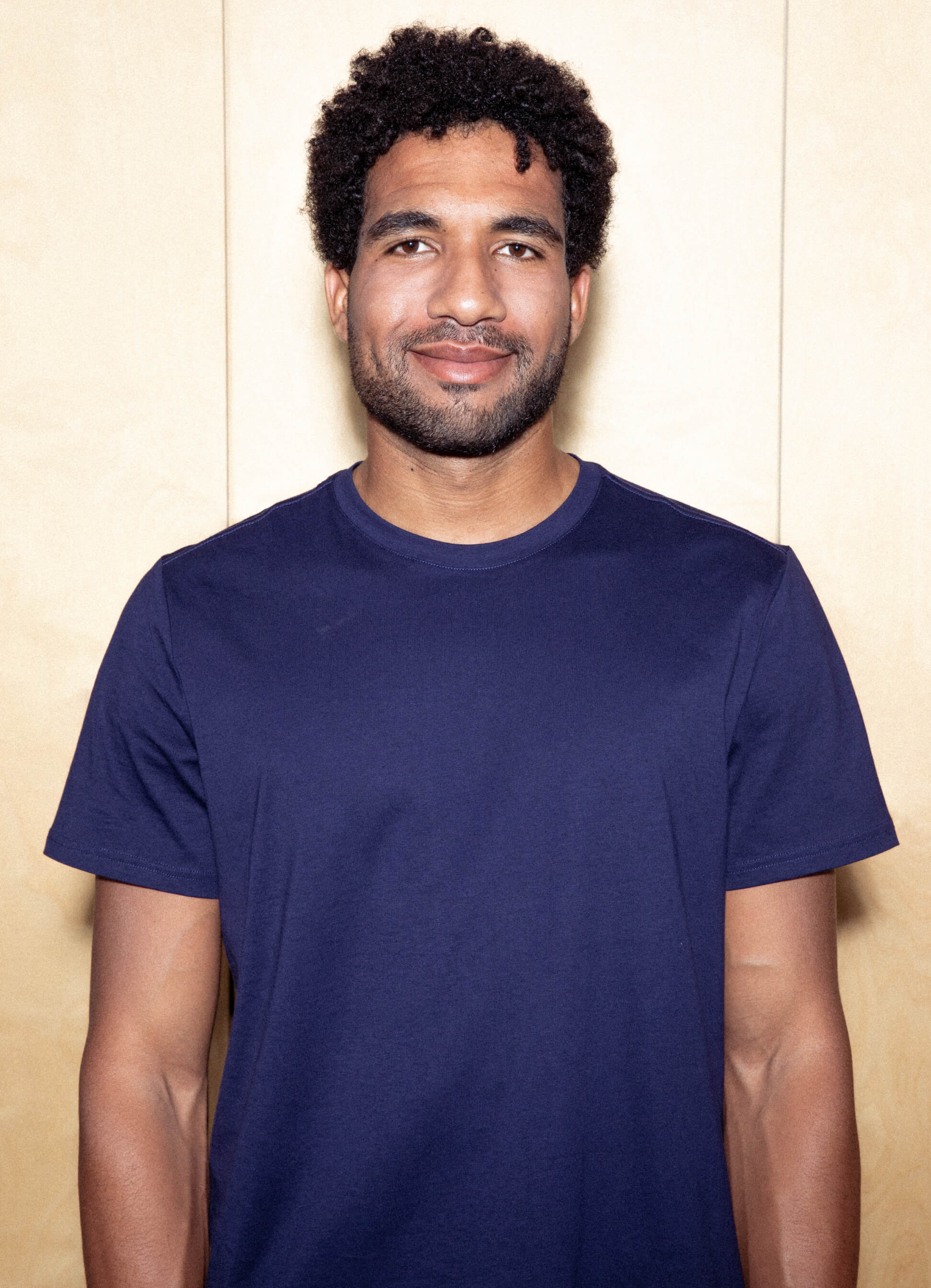 Retrato de homem sorridente com cabelo escuro encaracolado e barba veste uma camiseta azul-marinho contra um fundo colorido claro