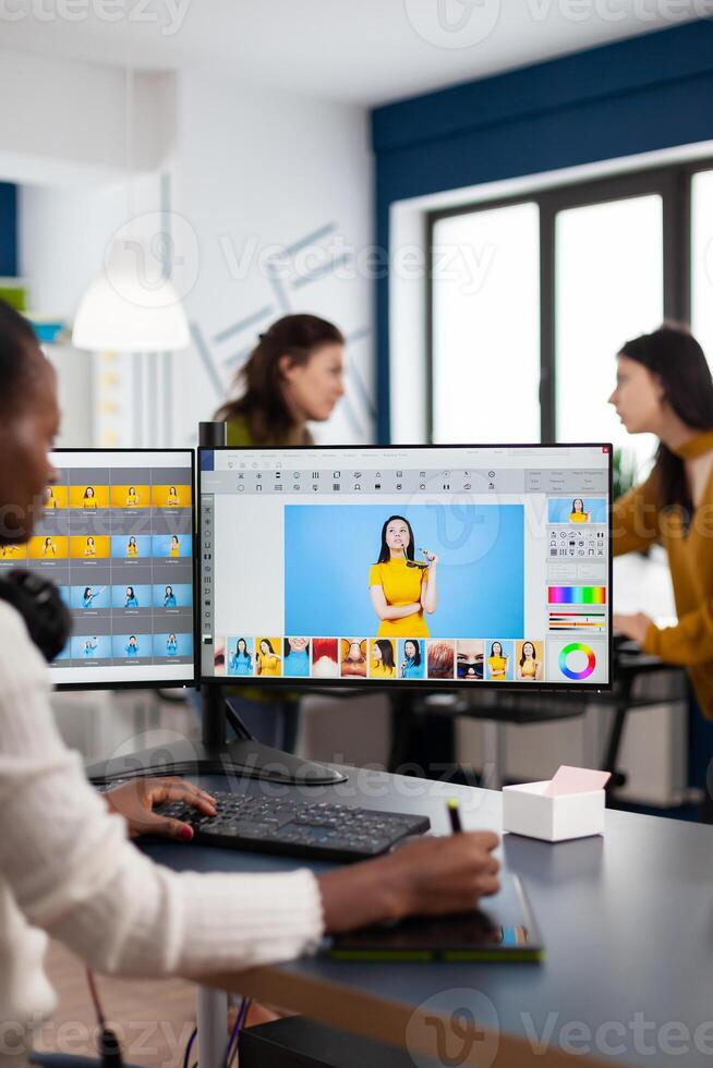 Black retoucher woman analysing images on pc with two monitors and retouching digital assets with stylus pencil. African project designer using photo editing software sitting in creative agency office