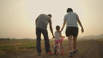 Asian father and mother taking their daughter pink bike in the meadow during the time the sunsets. Family concept. video