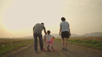 Asian father and mother taking their daughter pink bike in the meadow during the time the sunsets. Family concept. video