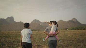 4k Video slow-motion, Daughter riding on father's back Mother standing nearby Coming out for a walk in the meadow Happily During the sunset. Family concept.