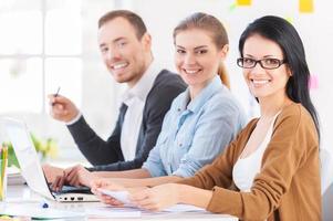 Coming up with fresh design solutions. Three cheerful young colleagues sitting together at their working place and smiling at camera photo