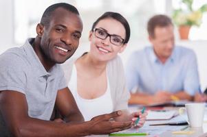 We are young and creative Two business people sitting together at the table and smiling while other people working on background photo