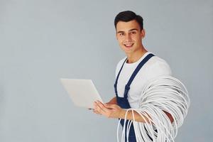 Uses laptop. Male worker in blue uniform standing inside of studio against white background photo