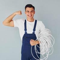 Holds cable. Male worker in blue uniform standing inside of studio against white background photo