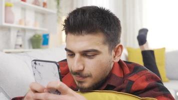 Happy young man holding mobile phone using smartphone device at home. Smiling young man subscribes to social media, buys online, orders products online in apps, addicted to phone. video