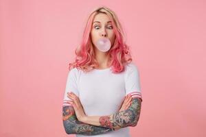 Young funny beautiful pink haired woman with tattooed hands, wears in white t-shirt, blows gum ball, squinted eyes on nose, stands over pink background with crossed arms. photo