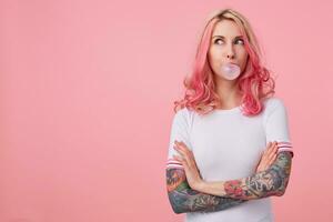 Portrait of young amazed beautiful pink haired lady with tattooed hands, wears in white t-shirt, blows gum ball, looking to the left in surprise, stands over pink background with copy space. photo