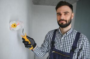 Young smiling professional worker in blue uniform standing with paint roller in new apartment for repairing over grey walls background, copy space. photo