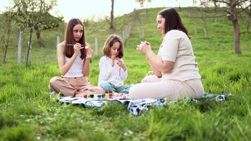 a family of three girls of different ages and European appearance are sitting in the garden on the green grass and are engaged in painting Easter eggs. Easter traditions. Spring holiday. video