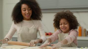 Cheerful African American mother woman teach little daughter child kid girl make flattening dough with rolling-pins in kitchen happy family use ecological products ingredients cooking together smiling video