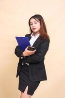 Young beautiful formal Asian executive woman in suit elegant standing and pose hugging clipboard and smartphone gadget to camera on yellow background for commercial, vacancy and business photo