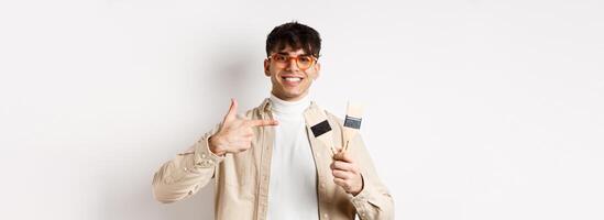 Hobbies and leisure concept. Cheerful young man in glasses pointing at painting brushes, renovating apartment, standing on white background photo